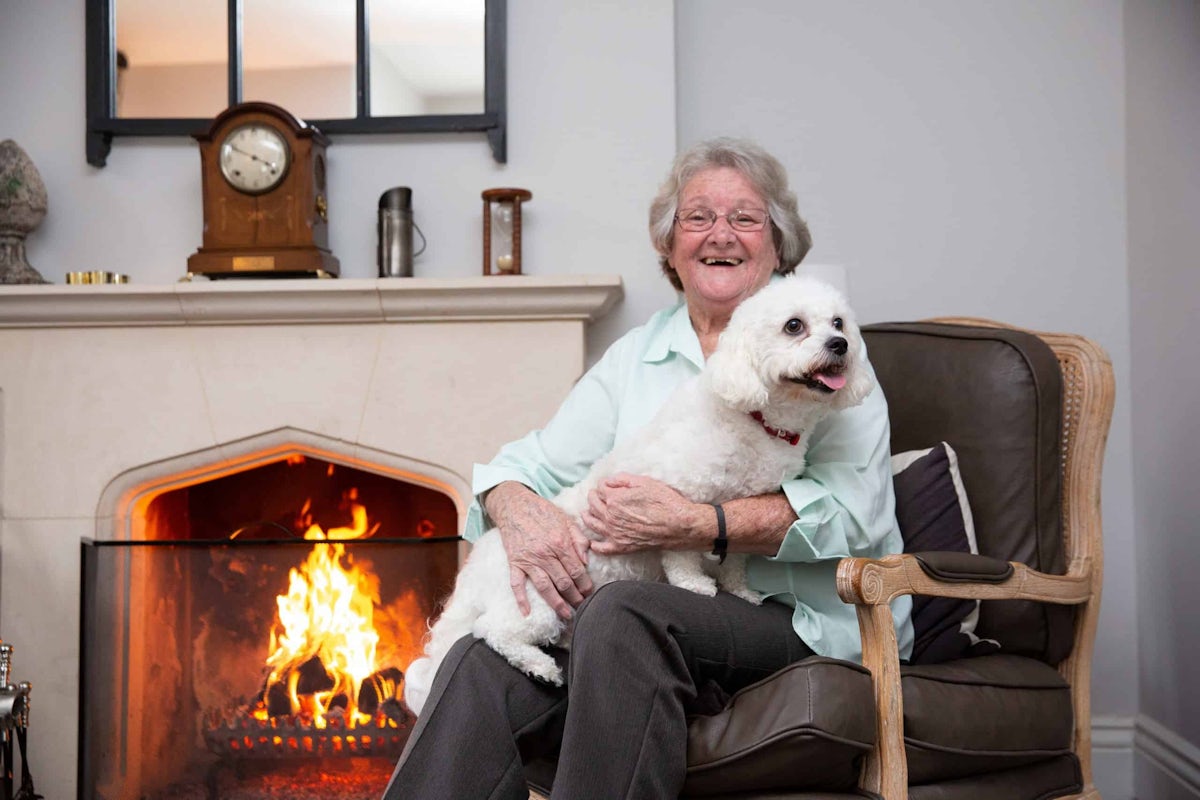 A woman enjoying live-in care with her white dog sitting on her lap by an open fire